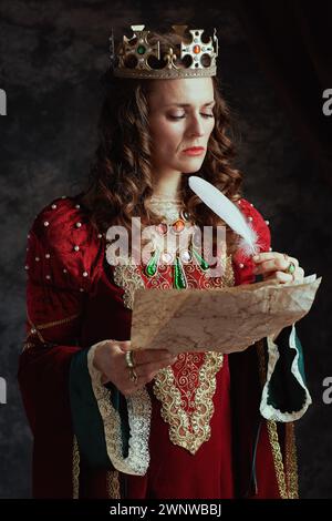 Nachdenkliche mittelalterliche Königin in rotem Kleid mit Pergament und Krone auf dunkelgrauem Hintergrund. Stockfoto