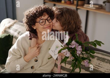 Süßer Junge, der seine Mutter auf die Wange küsst und sie umschließt, während er sich über die Couch beugt und ihr zu den Frühlingsferien gratuliert Stockfoto