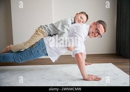 Ein energiegeladener Vater und sein kleiner Sohn trainieren zu Hause. Der Junge kletterte Dad auf den Rücken, während er Plank machte. Junge Familie, die zu Hause Aktivitätsspiele genießt. Stockfoto