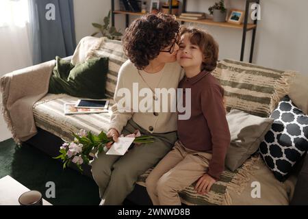 Liebevolle Mutter mit einem Haufen Blumen und einer Postkarte, die ihren liebenswerten Sohn umschließt und ihn auf die Wange küsst, während sie auf der Couch sitzt Stockfoto