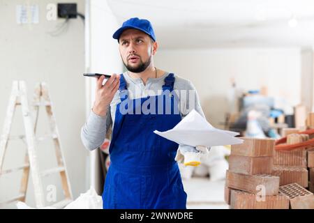 Fokussierter bärtiger Auftragnehmer in Blau, der insgesamt auf der Baustelle in Innenräumen stand und den Arbeitsprozess telefonisch diskutierte Stockfoto