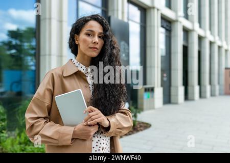 Eine selbstbewusste, lockige professionelle Frau steht vor einem modernen Bürogebäude, hält ein Tablet und sieht nachdenklich aus. Sie scheint konzentriert und bereit für das Geschäft in einem stilvollen Outfit zu sein. Stockfoto