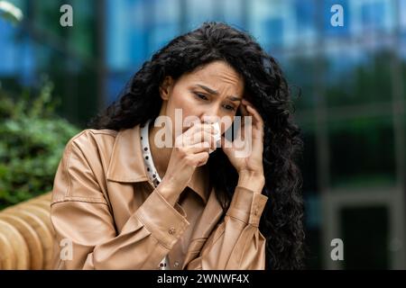 Eine beunruhigte Geschäftsfrau in professioneller Kleidung sitzt draußen und wischt mit einem Taschentuch Tränen ab, ein Blick von Traurigkeit und Sorge auf ihrem Gesicht vor einem städtischen Hintergrund. Stockfoto