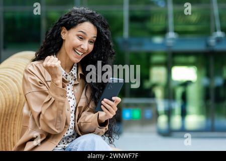 Junge Frau in legeren Geschäftskleidung, die im Freien sitzt und im Hintergrund ein modernes Gebäude hat, das Konnektivität und urbanen Lebensstil symbolisiert. Stockfoto