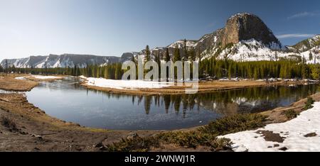 Dieses Panoramabild zeigt einen ruhigen Fluss, der sich durch eine malerische schneebedeckte Berglandschaft unter einem klaren blauen Himmel schlängelt Stockfoto