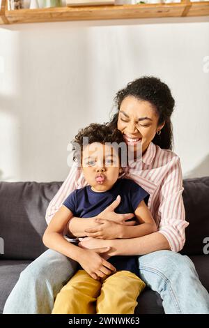 Fröhliche afroamerikanerin, die ihren kleinen, entzückenden Sohn hält, der vor der Kamera die Zunge ausstreckt Stockfoto