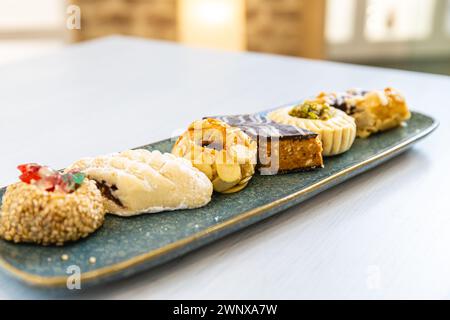 Horizontales Foto eine elegante Auswahl gemischter arabischer Süßigkeiten mit verschiedenen Aromen und Texturen, kunstvoll auf einer strukturierten blauen Servierplatte angeordnet. Stockfoto