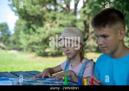 Ein Junge in einem blauen Hemd und ein Mädchen in einem Strohhut konzentrierten sich auf ein Brettspiel in einer natürlichen Umgebung. Dieses Bild fängt Kindheit, Freizeit und lea perfekt ein Stockfoto