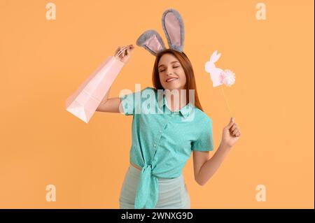 Junge Frau in Hasenohren mit Einkaufstasche und Papierkaninchen auf orangem Hintergrund. Osterangebot Stockfoto