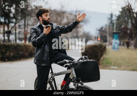 Eleganter Geschäftsmann auf dem Fahrrad in der Stadt, der ein Taxi ruft und gleichzeitig ein Smartphone hält Stockfoto