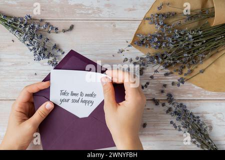 NEHMEN SIE SICH DIE ZEIT, UM IHRE SEELE GLÜCKLICH ZU MACHEN, indem Sie auf einer unterstützenden Nachricht auf Papier Notizen aus dem grünen Umschlag erinnern. Flache Zusammensetzung trockene Lavendelblüten. Konzept von innerem Glück, Verlangsamung digitaler Detox persönliche Erfüllung. Draufsicht Stockfoto