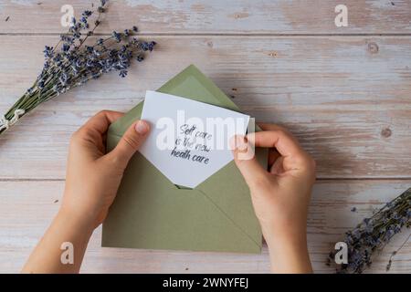 SELF CARE IST DER NEUE GESUNDHEITSTEXT auf unterstützender Nachricht Papier Notiz Erinnerung aus grünem Umschlag. Flache Zusammensetzung trockene Lavendelblüten. Konzept von innerem Glück, Verlangsamung digitaler Detox persönliche Erfüllung. Draufsicht Stockfoto