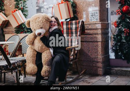 Lächelnde Frau im Wintermantel umarmt einen riesigen Teddybären in der Nähe einer Weihnachtskulisse, die ein Gefühl von Wärme und Fröhlichkeit weckt. Stockfoto