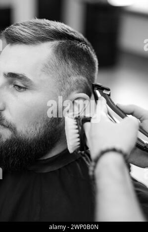 Barber rasiert die Kontur der ovalen Linie mit einem Haarschneider am Kopf des Kunden. Ein Mann mit Bart bekommt einen Haarschnitt in einem Friseurstuhl. Stockfoto