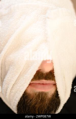 Ein Kunde liegt auf einem Stuhl in einem Friseurladen mit seinem Bart in ein Handtuch gewickelt. Den Bart vor der königlichen Rasur dämpfen. Stockfoto