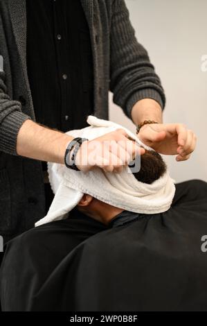 Der Friseur bedeckt das Gesicht des Kunden mit einem feuchten Handtuch, um den Bart abzudampfen. Stockfoto