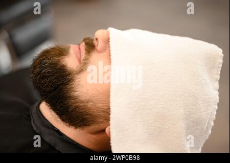 Ein Kunde liegt auf einem Stuhl in einem Friseurladen mit seinem Bart in ein Handtuch gewickelt. Ein Handtuch bedeckt die Augen des Mannes. Stockfoto