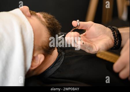 Ein Friseur beschneidet den Bart eines Kaukasiers, dessen Gesicht mit einem Handtuch bedeckt ist, mit einer Schere Stockfoto