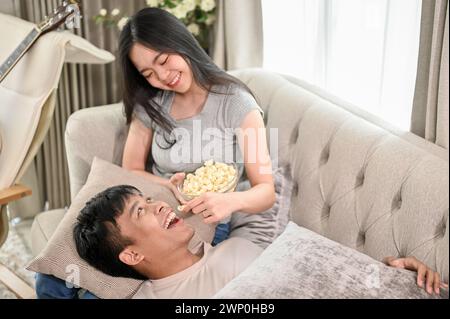 Ein glücklicher junger asiatischer Mann liegt auf dem Schoß seiner Freundin auf einem Sofa und teilt einen süßen und romantischen Moment zu Hause zusammen. Paare, verheiratetes Paar, Boyfr Stockfoto