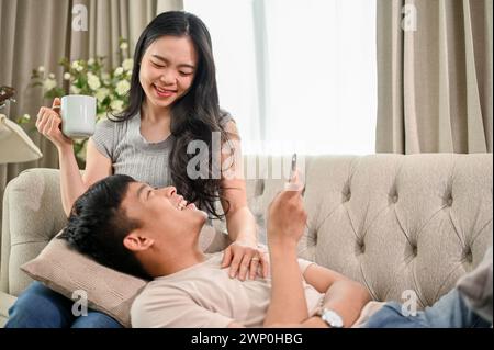 Ein glückliches junges asiatisches Paar genießt es, sich auf einem Sofa im Wohnzimmer zu unterhalten und zu entspannen, die Freundin trinkt Kaffee, während ihr Freund li Stockfoto