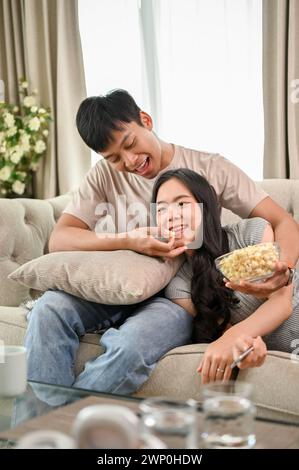 Ein liebenswerter, junger asiatischer Freund füttert Popcorn an seine Freundin, während er sich entspannt und zusammen einen Film auf dem Sofa ansieht. Lover und dom Stockfoto