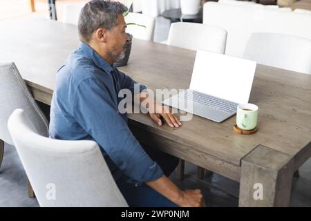 Biracial Senior man arbeitet an einem Laptop an einem modernen Esstisch mit Kopierraum Stockfoto