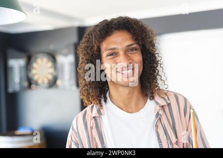 Ein junger birassischer Mann mit lockigen Haaren lächelt warm und trägt ein gestreiftes Hemd über einem weißen T-Shirt Stockfoto