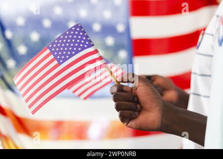 Die Hand eines afroamerikanischen Mannes, der eine kleine amerikanische Flagge hält, mit einer größeren Flagge im Hintergrund Stockfoto