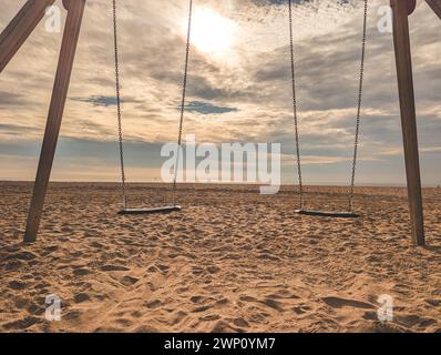 Schaukeln hängen an aufgerichteten Holzpfählen am Sandstrand mit Ketten bei sonnigem Tageslicht an Wolken, die tagsüber die helle Sonne am blauen Himmel bedecken Stockfoto