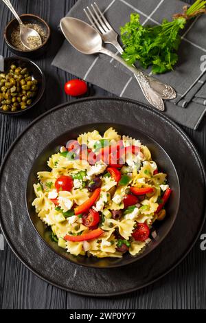 Farfalle Salat, Bow Tie Pasta Salat mit kalamata und grünen Oliven, Paprika, Kapern, Zwiebeln, Gurken, Tomaten und Feta in schwarzer Schüssel auf schwarzem Woo Stockfoto