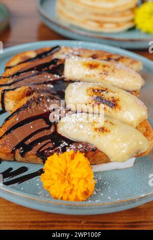 Ein Teller Bananenbrot mit Schokoladensoße und Puderzucker. Der Teller steht auf einem Tisch mit einer Vase voller Blumen im Hintergrund Stockfoto