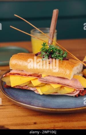 Ein Sandwich mit Schinken, Käse und Gurken steht auf einem blauen Teller. Ein Glas Orangensaft steht neben dem Sandwich Stockfoto