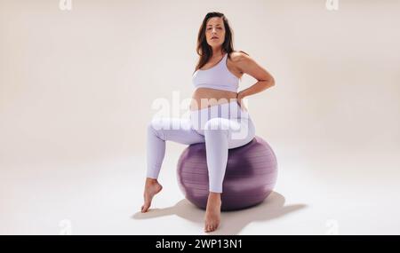 Junge Frau übt Schwangerschaftsyoga auf einem Stabilitätsball in einem Studio. Sie trägt Fitnesskleidung und zeigt ihr Baby mit Kraft und Flexi Stockfoto