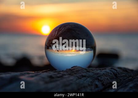 Glaskugel auf einem Zweig bei Sonnenuntergang am Strand, das Meer und die untergehende Sonne spiegeln sich in der Kugel Stockfoto