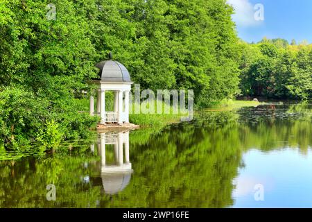 Weißer hölzerner Pavillon am Fluss, umgeben von Bäumen. Blick an einem ruhigen Sommermorgen mit klarem Himmel und Reflexionen auf dem Wasser. Stockfoto