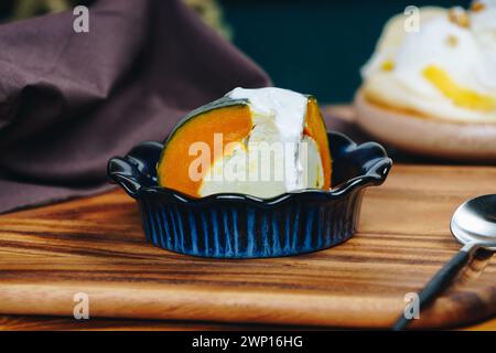 Eine blaue Schüssel mit einem Stück Kürbiskuchen steht auf einem Holztisch. Der Kuchen wird mit Schlagsahne belegt und hat einen Kürbis darauf. Die Schüssel wird auf einen gelegt Stockfoto