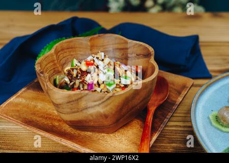 Auf einem Holzteller befindet sich eine Schüssel mit einem Löffel daneben. Die Schüssel ist mit einer Vielzahl von Zutaten gefüllt, darunter Nüsse, Gemüse und Obst. Stockfoto