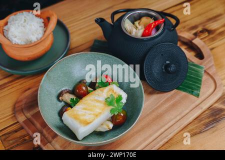 Ein Teller mit Fisch und Gemüse sitzt auf einem Holzbrett. Eine Teekanne ist ebenfalls auf dem Brett. Die Szene ist warm und einladend, mit dem Essen und der Teekanne Stockfoto