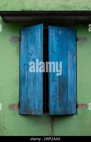 Zwei verblasste, abgebrochene, lackierte, leicht offene blaue Holzläden an einer grünen Wand an einem Haus im ländlichen Nordvietnam. Stockfoto