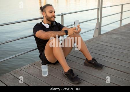 Der Typ in Sportswear sitzt draußen und überprüft Fitness-Apps auf dem Smartphone Stockfoto