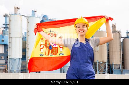 Junge Ingenieurin im Helm, die Landesflagge von Spanien schwenkt, während sie vor großen Tanks in der Chemiefabrik steht Stockfoto