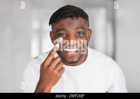 Der Kopfschuss des fröhlichen afrikanischen Mannes trägt Gesichtscreme auf die Wange auf Stockfoto
