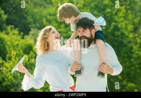 Kinder spielen mit einfachen Papierflugzeugen an sonnigen Tagen. Gemeinsame Familienzeit. Porträt des glücklichen Vaters, der Sohn Huckepack auf seinen Schultern reitet und Stockfoto