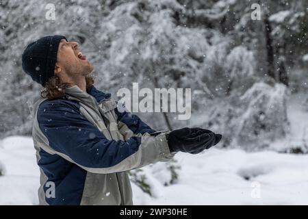 Ein Mann in einer blau-grauen Jacke wirft Schnee in die Luft. Er trägt einen schwarzen Hut und Handschuhe. Vorstellung von Freude und Verspieltheit, wie der Mann es genießt Stockfoto