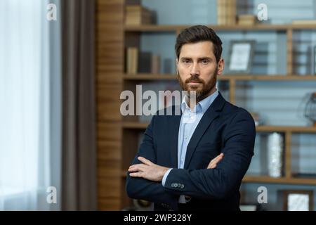 Ein ernster junger Mann in einem Business-Anzug steht im Büro und blickt selbstbewusst in die Kamera, indem er seine Arme über der Brust kreuzt. Stockfoto