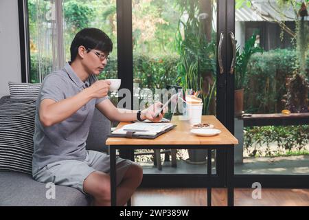 Junger asiatischer Mann hält eine Tasse Kaffee und mit Smartphone, während er in der Nähe des Fensters im Heimbüro sitzt. Stockfoto