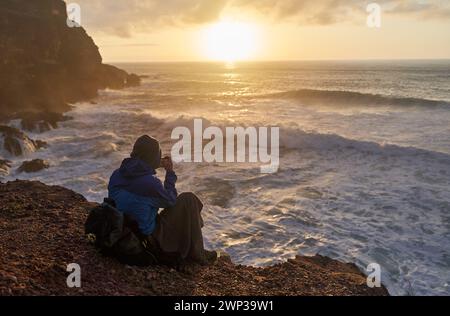 Die Person sitzt auf einer Klippe und blickt auf das Meer, während die Sonne über Wasser, Himmel und Horizont untergeht Stockfoto