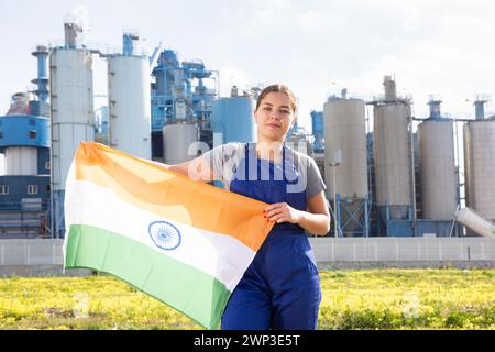 Vor dem Hintergrund der Fabrik außerhalb der Stadt steht ein ausgewogenes Mädchen im Overall mit indischer Flagge Stockfoto