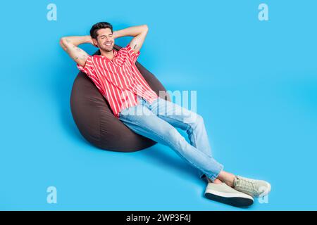Ganzkörperfoto des friedlichen entspannten Guy tragen stilvolle Hemd Jeans liegen auf Pouf Palmen hinter Kopf Nehmen Sie ein Nickerchen isoliert auf blauem Hintergrund Stockfoto