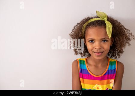 Ein junges schwarzes Mädchen mit lockigen Haaren, ein Regenbogengestreiftes Tanktop und ein grünes Stirnband, isoliert auf weißem Hintergrund, glücklich, lächelnd. Minimalistisch und Stockfoto